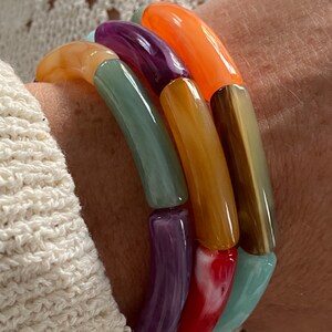 Peut inclure: Un ensemble de bracelets colorés en perles de plastique lisses et arrondies. Les bracelets sont empilés sur le poignet d'une personne et présentent des nuances d'orange, de violet, de vert, de jaune et de rouge.