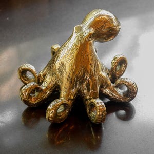 Puede incluir: Una detallada escultura de pulpo de lat&oacute;n con ocho tent&aacute;culos rizados. El cefal&oacute;podo de color dorado tiene una superficie texturizada y se muestra sobre una superficie oscura y reflectante. La escultura es un acento n&aacute;utico decorativo.