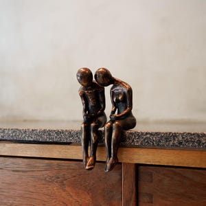 Puede incluir: Una escultura de bronce de dos figuras sentadas una al lado de la otra, con una figura inclinada hacia la otra. La escultura está colocada sobre una encimera de granito encima de un mueble de madera. Las figuras son detalladas y tienen un acabado liso y pulido.