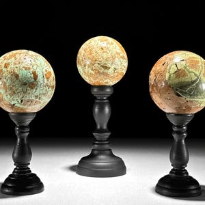 Può includere: Tre sfere in pietra levigata con motivi maculati nei toni del marrone chiaro, arancione e verde chiaro, ciascuna poggiata su un piedistallo in legno nero. Le sfere presentano venature naturali e variazioni di colore sulle superfici.