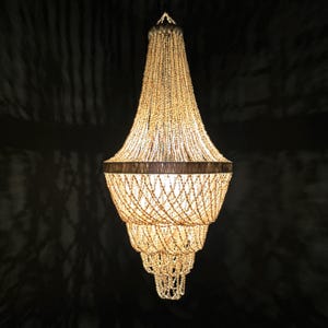 Puede incluir: Un candelabro en capas con un diseño de cuentas. La lámpara tiene una parte superior cónica y una base más ancha en capas. Las cuentas son de color beige claro y la lámpara cuelga de un solo punto. El fondo es oscuro.