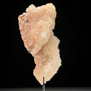May include: A peach-colored mineral specimen with a crystalline structure. The mineral is mounted on a black metal stand against a black background. The crystals have a rough, textured appearance, with variations in color from light peach to a darker, reddish-brown hue.