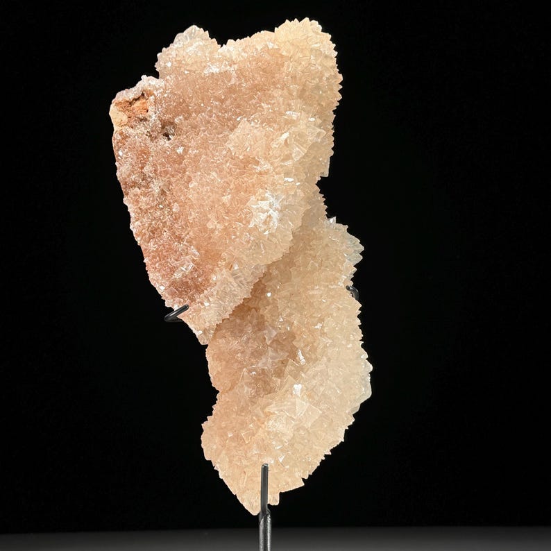 May include: A peach-colored mineral specimen with a crystalline structure. The mineral is mounted on a black metal stand against a black background. The crystals have a rough, textured appearance, with variations in color from light peach to a darker, reddish-brown hue.