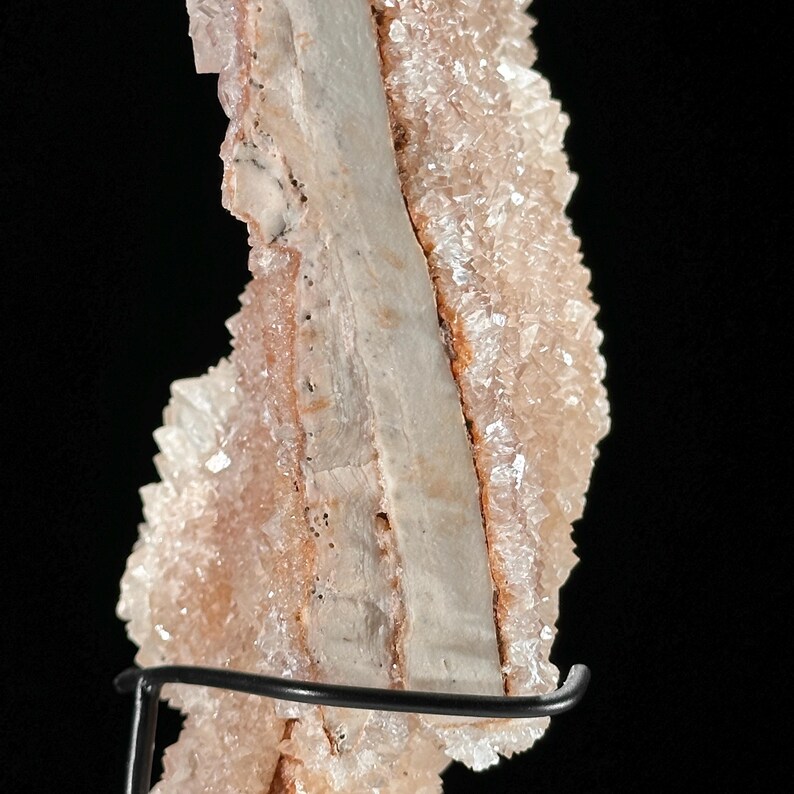 May include: Close-up of a natural mineral specimen. The stone features a central, light-colored band flanked by layers of crystalline formations in shades of peach and cream. The mineral is mounted on a black metal stand against a black background.