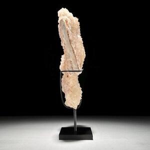 May include: A light peach-colored crystal formation with a central, lighter-toned section, mounted on a black metal stand. The crystal has a textured, rough surface. The stand has a square base and a curved support.