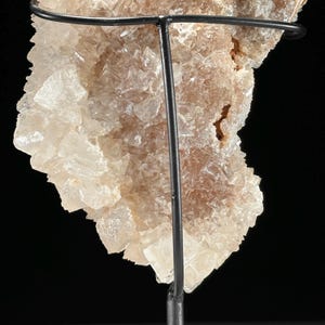 May include: A close-up of a mineral specimen, showcasing a cluster of crystalline structures in shades of beige and off-white. The mineral is mounted on a black metal stand, with the background a solid black. The crystals have a rough, textured appearance.