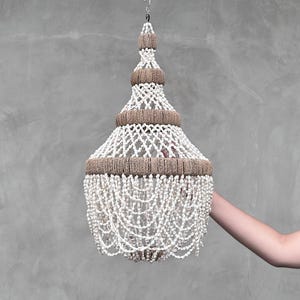 May include: A decorative chandelier with a tiered design, suspended from a chain. It features a white, beaded body and brown woven trim. The chandelier hangs against a grey background. The design is bohemian and suitable for interior decor.