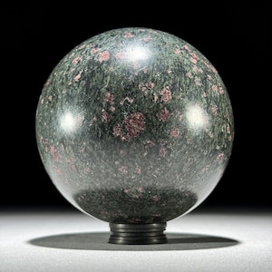 May include: A polished, spherical decorative object. The sphere is a deep green color with small, irregular red inclusions. It sits on a dark, circular base. The overall effect is of a natural stone with a smooth, reflective surface.