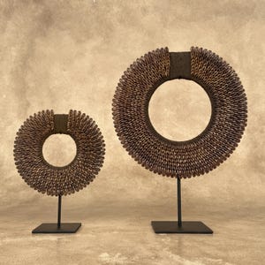 Puede incluir: Dos esculturas circulares decorativas. Las esculturas están hechas de pequeñas conchas marrones dispuestas en forma de anillo. Cada anillo está montado sobre un soporte de metal negro con una base cuadrada. El fondo es de color beige neutro.