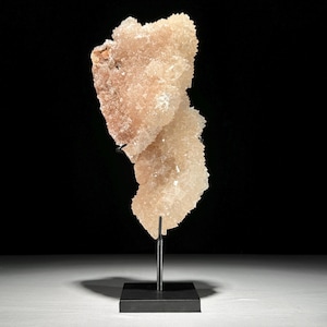 May include: A peach-colored crystal cluster on a black metal stand. The crystal formation has a rough, textured surface and is mounted on a thin, vertical rod attached to a square base. The background is black.