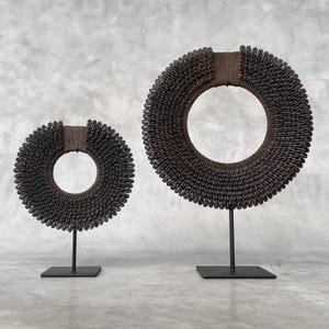 May include: Two decorative, dark brown, circular sculptures. Each sculpture is composed of small, dark beads arranged in a ring shape. The rings are mounted on black metal stands with square bases. The sculptures have a rustic, earthy aesthetic.