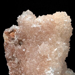 May include: Close-up of a mineral specimen with a crystalline structure. The mineral features a pale peach color with translucent, angular crystals. The background is solid black, highlighting the intricate details of the natural formation.