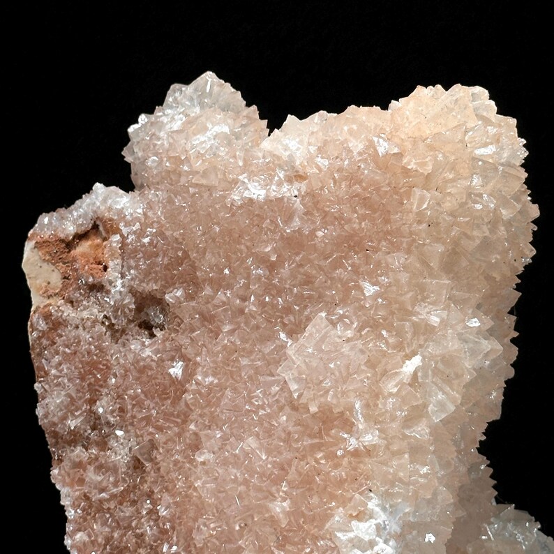 May include: Close-up of a mineral specimen with a crystalline structure. The mineral features a pale peach color with translucent, angular crystals. The background is solid black, highlighting the intricate details of the natural formation.