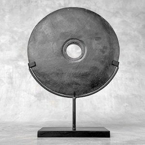 Pode incluir: Uma escultura circular em pedra cinza escuro com um buraco central, montada em um suporte de metal preto. A pedra tem uma superfície texturizada e é exibida em um fundo neutro. O suporte tem uma base retangular.