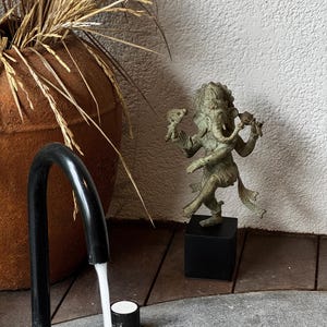Könnte beinhalten: Eine bronzefarbene Statue der hinduistischen Gottheit Ganesha, die auf einem schwarzen quadratischen Sockel steht. Die Statue befindet sich in einer dynamischen Pose mit mehreren Armen und einem Elefantenkopf. Ein großer, rostfarbener Topf mit getrockneten Gräsern befindet sich im Hintergrund.