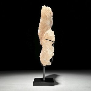 May include: A large, light peach-colored crystal formation on a black metal stand. The crystal has a rough, textured surface and is displayed against a black background. The stand has a square base and a curved support.