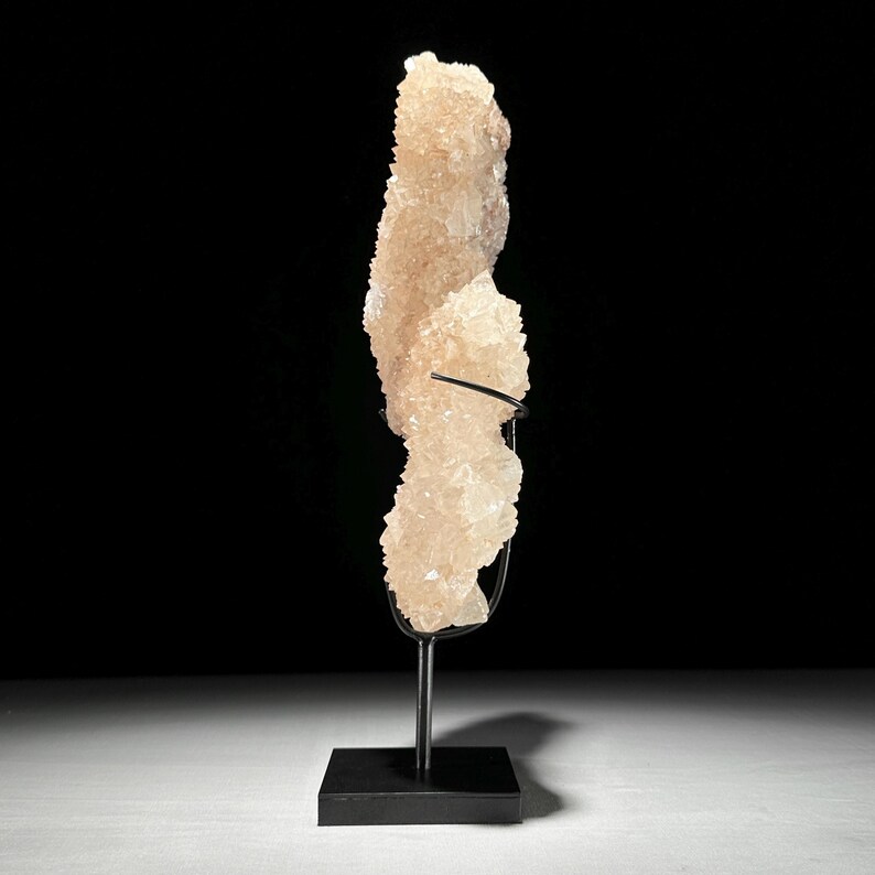 May include: A large, light peach-colored crystal formation on a black metal stand. The crystal has a rough, textured surface and is displayed against a black background. The stand has a square base and a curved support.