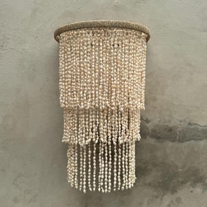 May include: A three-tiered wall hanging light fixture featuring a circular frame wrapped in natural fibre, adorned with numerous strands of small, off-white seashells that cascade downward in a layered, fringe-like arrangement.