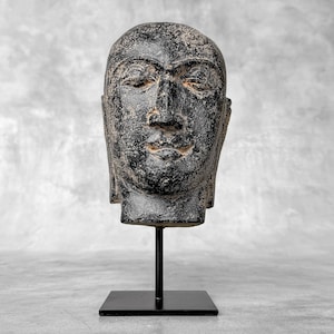 Peut inclure: Une sculpture en pierre noire patinée représentant un visage serein, montée sur un support en métal noir. La sculpture présente des yeux fermés, un nez bien défini et une bouche légèrement entrouverte. La tête est exposée sur une base carrée.