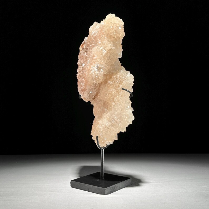 May include: A light peach-colored crystal cluster on a black metal stand. The crystal formation has a rough, textured surface and is mounted on a square base. The background is black, highlighting the crystal's unique shape.