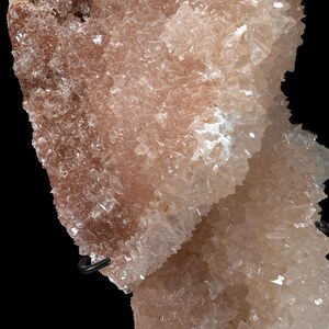 May include: Close-up of a mineral specimen with a crystalline structure. The mineral has a pale pink hue with areas of white and brown. The crystals are small and densely packed, creating a textured appearance. The background is black.