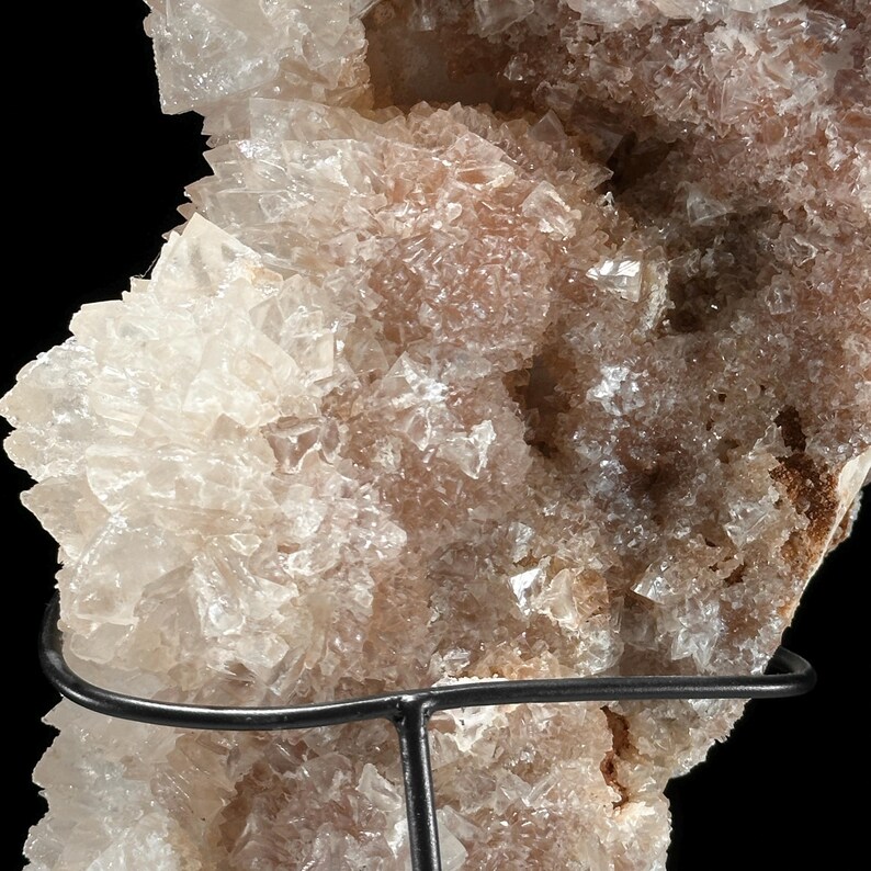 May include: Close-up of a mineral specimen with a crystalline structure. The crystals are primarily off-white and light brown, with a sparkling, reflective quality. The mineral is mounted on a black metal stand against a black background.