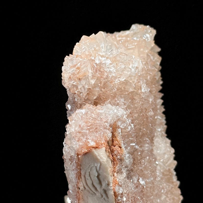 May include: Close-up of a mineral specimen with a crystalline structure. The mineral has a pale peach color with areas of white and a darker, reddish-brown hue. The background is solid black, highlighting the mineral's texture and form.