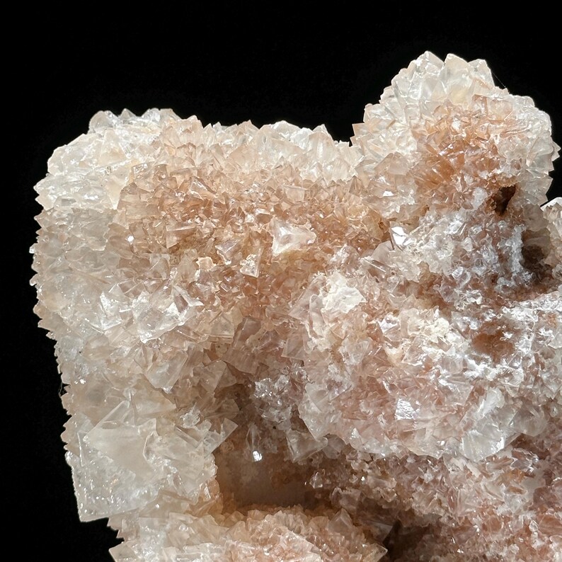 May include: Close-up of a crystalline mineral formation. The mineral features a cluster of translucent, light-colored crystals with hints of peach and brown hues. The crystals have sharp, angular shapes, creating a textured surface against a black background.