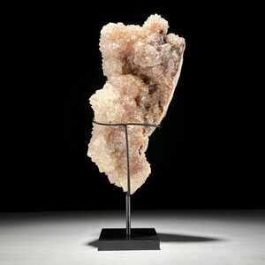 May include: A large, light-colored crystal cluster with a rough, textured surface. The crystal is mounted on a black metal stand with a square base, set against a dark background. The crystal's color ranges from off-white to light brown.
