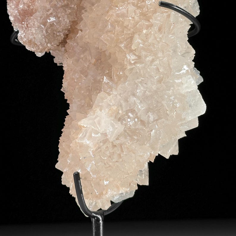 May include: A light peach-colored mineral specimen with a crystalline structure, mounted on a black metal stand. The mineral has a rough, textured surface with many small, pointed crystals. The background is black.