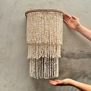 Op de afbeelding: Een wandlamp met drie niveaus, voorzien van een cirkelvormig frame omwikkeld met natuurlijke vezels, versierd met talloze strengen kleine, gebroken witte schelpen die verticaal hangen voor een trapsgewijze, gelaagde en textuurrijke cilindrische uitstraling.