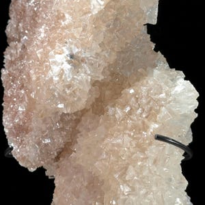 May include: Close-up of a pale pink mineral formation with a crystalline structure. The mineral has a rough, textured surface with many small, individual crystal formations. The background is black, highlighting the mineral's color and texture.