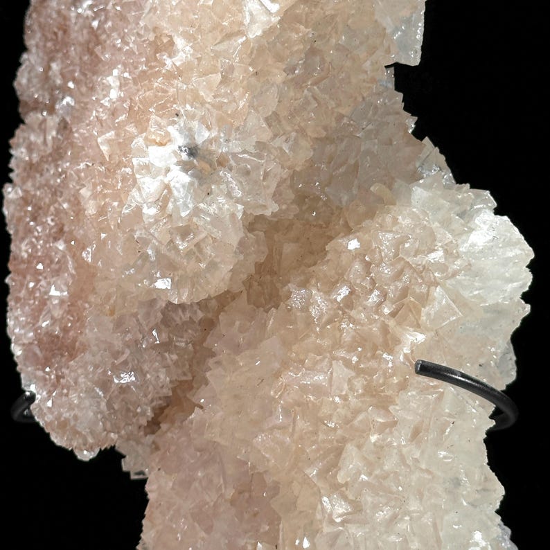 May include: Close-up of a pale pink mineral formation with a crystalline structure. The mineral has a rough, textured surface with many small, individual crystal formations. The background is black, highlighting the mineral's color and texture.