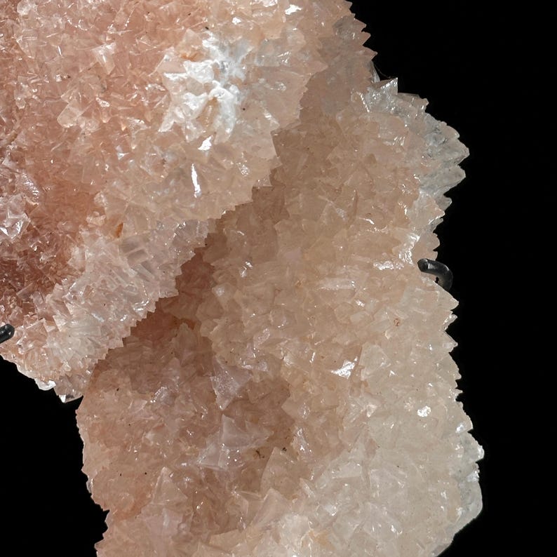 May include: Close-up of a mineral formation with a crystalline structure. The mineral has a pale peach color and a rough, textured surface. The crystals are small and densely packed, creating a layered appearance against a black background.