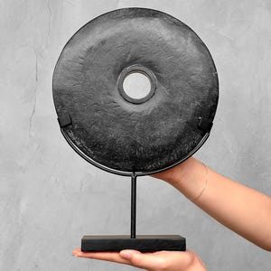 Op de afbeelding: Een zwart, cirkelvormig decoratief object met een centraal gat, gemonteerd op een zwarte metalen standaard. Het object heeft een textuuroppervlak en wordt gepresenteerd tegen een grijze achtergrond. De standaard heeft een rechthoekige basis.