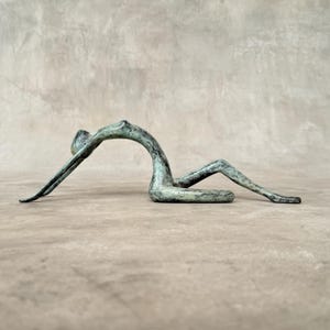 Puede incluir: Escultura abstracta de bronce de una persona en posición reclinada. La escultura tiene un acabado de pátina verde.