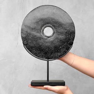 May include: A black, circular decorative object with a central hole, mounted on a black metal stand and base. The textured surface of the disc has a speckled appearance. The stand is held by a person.