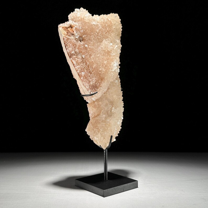 May include: A large, light peach-colored crystal formation with a rough, textured surface. The crystal is mounted on a black metal stand with a square base, showcasing its natural, irregular shape. The background is black.