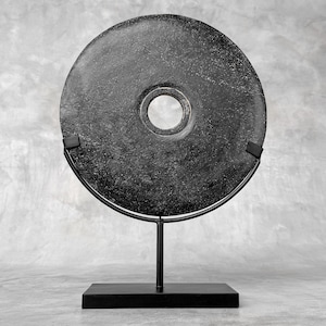 Könnte beinhalten: Eine kreisförmige schwarze Steinscheibe mit einem zentralen Loch, montiert auf einem schwarzen Metallständer. Die Scheibe hat eine texturierte, verwitterte Oberfläche mit feinen grauen Sprenkeln. Der Ständer besteht aus einem rechteckigen Fuß.
