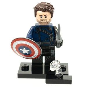 Pu&ograve; includere: Una minifigure in plastica con capelli castani e barba, che indossa una giacca blu scuro e pantaloni neri. La figura tiene uno scudo circolare rosso, bianco e blu in una mano e una piccola spada grigia nell'altra, su una base nera.