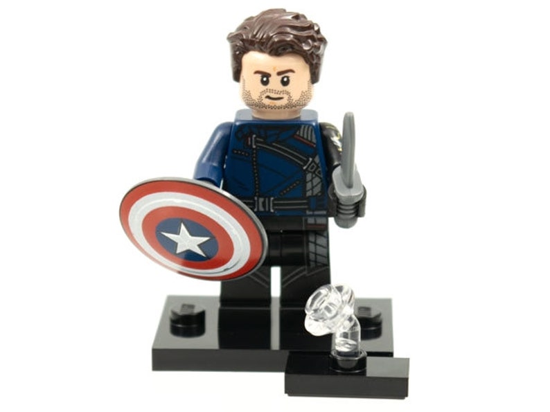 Pu&ograve; includere: Una minifigure in plastica con capelli castani e barba, che indossa una giacca blu scuro e pantaloni neri. La figura tiene uno scudo circolare rosso, bianco e blu in una mano e una piccola spada grigia nell'altra, su una base nera.