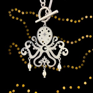 Puede incluir: Un collar con un colgante de plata en forma de pulpo con un diseño de estrella y media luna. El colgante está unido a un cierre de palanca de plata.