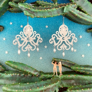 Puede incluir: Un par de pendientes de plata con un diseño de pulpo con una luna creciente y estrellas. Los pendientes tienen tres gotas de perlas a cada lado.