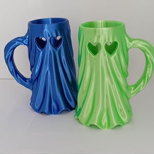 K&ouml;nnte beinhalten: Zwei Tassen in Geisterform mit drapierter Stofftextur. Eine Tasse ist metallisch blau, die andere metallisch hellgr&uuml;n. Jede Tasse hat zwei herzf&ouml;rmige Ausschnitte am oberen Rand und einen geschwungenen Henkel an der Seite.