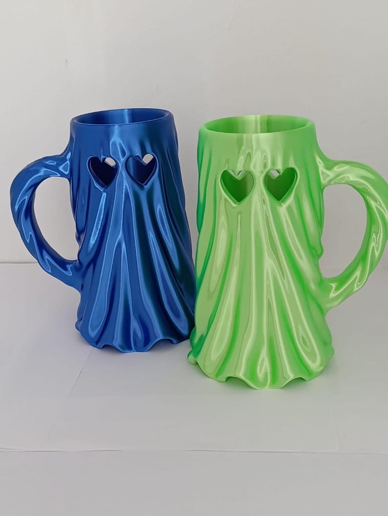 K&ouml;nnte beinhalten: Zwei Tassen in Geisterform mit drapierter Stofftextur. Eine Tasse ist metallisch blau, die andere metallisch hellgr&uuml;n. Jede Tasse hat zwei herzf&ouml;rmige Ausschnitte am oberen Rand und einen geschwungenen Henkel an der Seite.