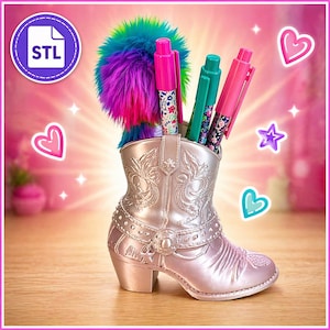 Könnte beinhalten: Ein silberfarbener Cowboy-Stiefel-Stifthalter mit dekorativen Blumen- und Sternakzenten. Der Stiefel enthält mehrere Stifte mit Blumen- und einfarbigen Designs sowie einen bunten, flauschigen Pompon. Der Hintergrund ist rosa mit Herz- und Sternformen.