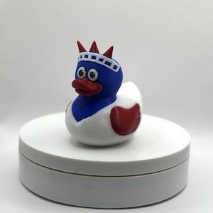 May include: A novelty rubber duck, coloured red, white, and blue, styled as the Statue of Liberty. The duck features a blue head, white body, and red accents, including a crown and heart-shaped wing. It rests on a white, circular platform.