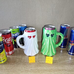 K&ouml;nnte beinhalten: Zwei geisterf&ouml;rmige Dosenhalter, einer wei&szlig; und einer gr&uuml;n, jeweils mit herzf&ouml;rmigen Augenausschnitten. Sie sind zwischen verschiedenen Soda- und Energy-Drink-Dosen platziert, mit gelben Etiketten f&uuml;r 330 ml und 355 ml Gr&ouml;&szlig;en.