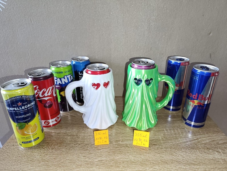 K&ouml;nnte beinhalten: Zwei geisterf&ouml;rmige Dosenhalter, einer wei&szlig; und einer gr&uuml;n, jeweils mit herzf&ouml;rmigen Augenausschnitten. Sie sind zwischen verschiedenen Soda- und Energy-Drink-Dosen platziert, mit gelben Etiketten f&uuml;r 330 ml und 355 ml Gr&ouml;&szlig;en.