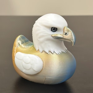May include: A 3D-printed decorative duck figurine with an eagle head. The eagle head is white, with a detailed beak and eye. The body transitions from gold to blue, with a white wing detail. A unique, patriotic home decor item.